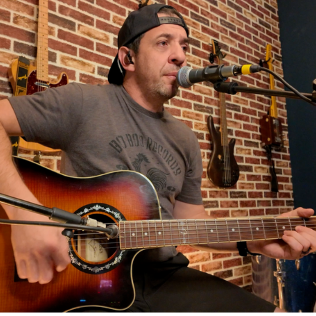 Sean Eddy playing live acoustic music in North Texas: acoustic guitar and vocals in front of a brick wall lined with guitars